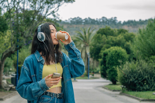 A relaxed young woman listening to music and drinking a takeaway beverage while walking down the street