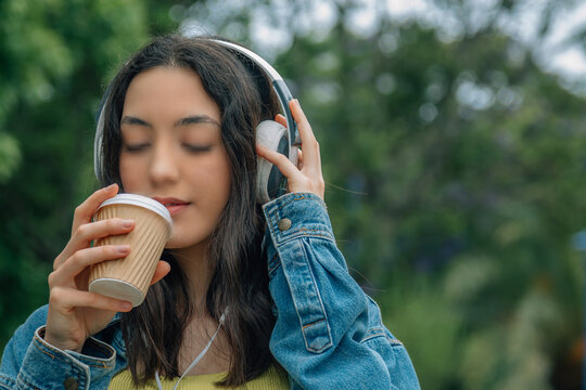 A relaxed young woman listening to music and drinking a takeaway beverage while walking down the street