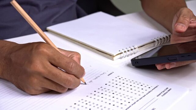 Close up of student writing essay while using smartphone for research