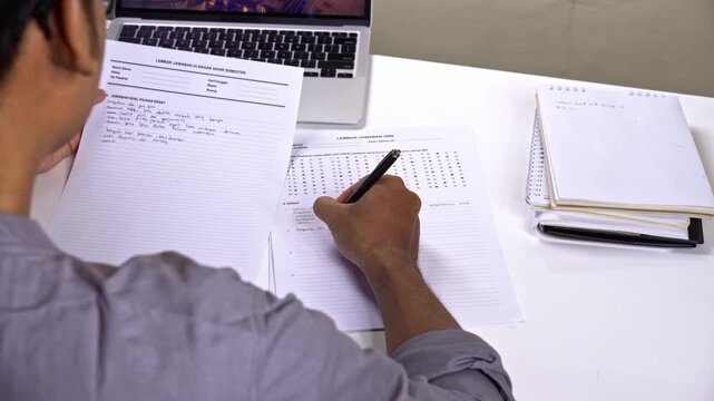 Over the shoulder shot of student taking exam and writing essay answer at desk