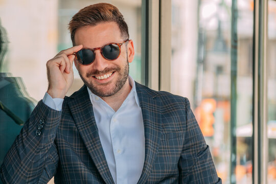 A confident businessman, elegantly dressed and wearing sunglasses, is standing in front of the office building.