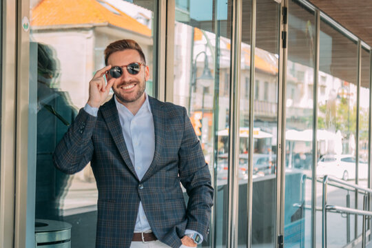 A confident businessman, elegantly dressed and wearing sunglasses, is standing in front of the office building.