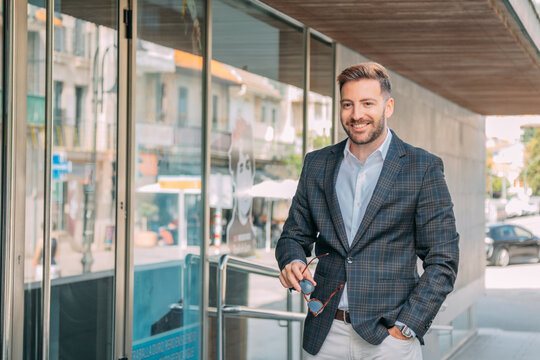 A confident businessman, elegantly dressed and wearing sunglasses, is standing in front of the office building.