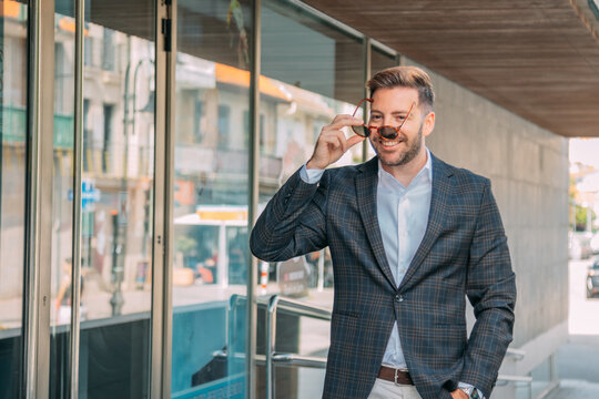 A confident businessman, elegantly dressed and wearing sunglasses, is standing in front of the office building.