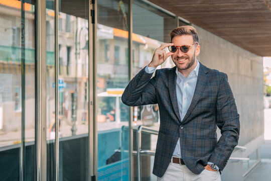 A confident businessman, elegantly dressed and wearing sunglasses, is standing in front of the office building.