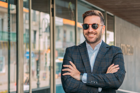 A confident businessman, elegantly dressed and wearing sunglasses, is standing in front of the office building.
