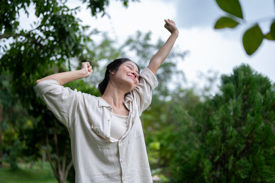 Asian woman stretching both arms upward joyfully in outdoor green garden.