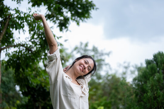 Asian woman stretching one arm up smiling joyfully in outdoor garden.