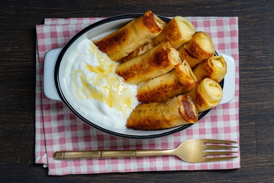 Delicious fried crepes filled with cottage cheese, served with sweet honey and sour cream in white ceramic dish on rustic checkered napkin. Perfect morning breakfast concept flat lay