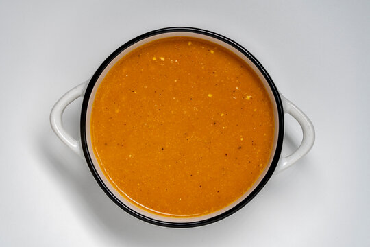 Top view of creamy pumpkin soup in white pot. Delicious hot orange cream soup in white ceramic pot with handles on clean white background. Minimalist food concept for autumn seasonal healthy eating