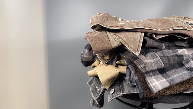 Stack of folded casual men's clothing featuring denim jackets and a plaid flannel shirt against a grey background with copy space.