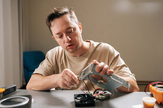 Focused young adult repairman fixing computer hardware components, examining circuit boards and connecting wires to diagnose issues and perform maintenance on electronic devices at workbench.