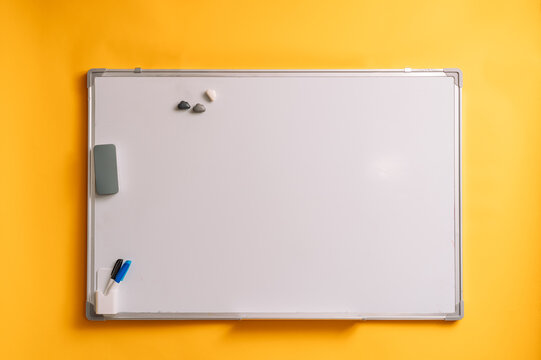 Blank whiteboard with accessories on a yellow wall in a gray frame.
