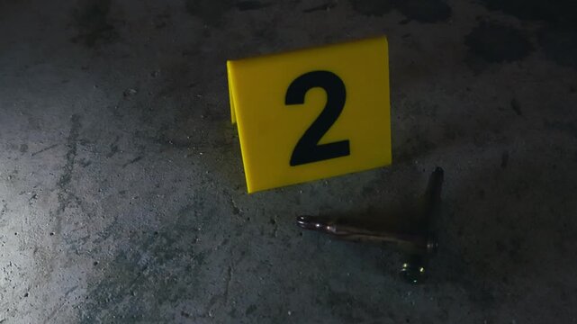 Yellow evidence marker with the number two next to spent high caliber bullet casings lying on a dusty concrete floor at a crime scene. Forensic investigation of a shooting in progress