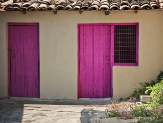 horizontal composition highlighting the striking contrast of a magenta wooden door, window mesh grid, terracotta tiles, and beige stucco in Kiato. © Dimitrios