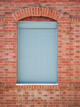 Industrial red brick wall with an arched window opening fitted with a blue-grey rolling shutter in Athens, Greece.