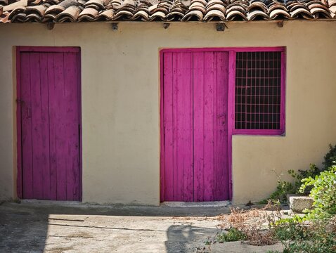 horizontal composition highlighting the striking contrast of a magenta wooden door, window mesh grid, terracotta tiles, and beige stucco in Kiato.