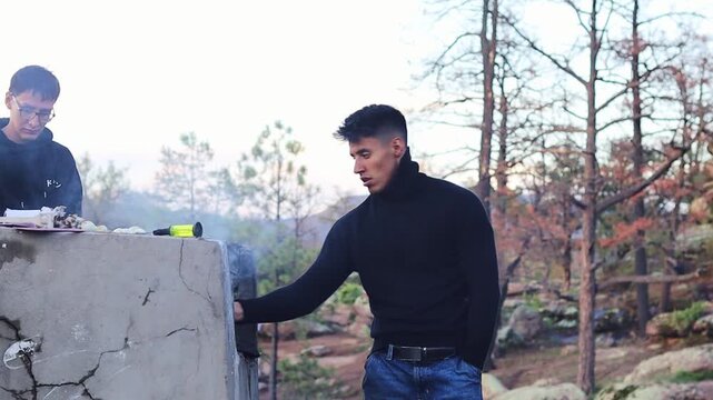 Young men cooking outdoors on a barbecue, stoking the fire with a poker and enjoying a relaxing picnic in the woods on a beautiful autumn afternoon. Slow motion video