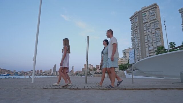 A group of four people strolls along the sandy beach. They move towards modern buildings while enjoying the view and the warm weather. The sky is clear and bright above them.