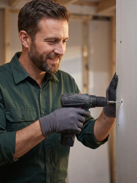 A smiling worker secures a drywall panel with a power drill during interior construction. Skilled installation work shows professional craftsmanship and progress in a building renovation