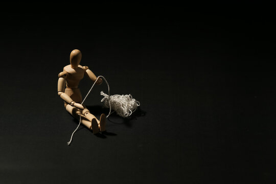 Wooden mannequin sitting on the floor trying to untangle a messy ball of white string. Concept of problem solving, complicated situation, confusion or brain.