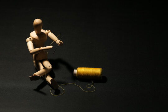 Wooden mannequin sitting on the floor sewing with a needle and a spool of yellow thread. Concept of self repair, recovery, healing, therapy or fixing problems.