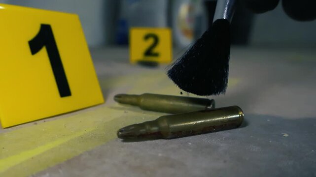 Forensic expert in gloves using a brush to dust for fingerprints on high caliber bullet casings at a crime scene, with yellow evidence markers visible on the ground for investigation