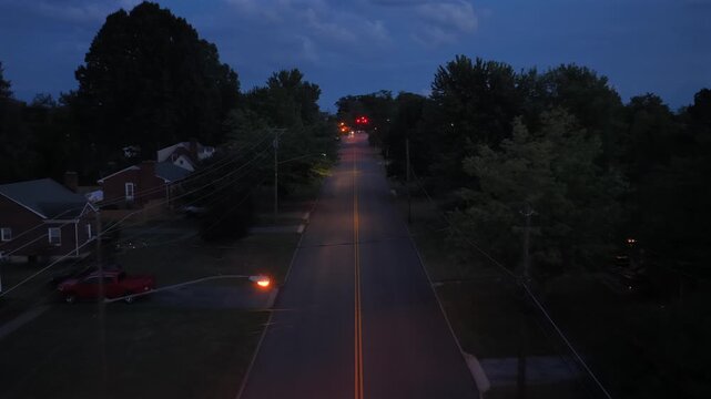 A drone flies along a Lynchburg street in Virginia at dusk with brick houses either side, an oncoming car and broken cloud still bright overhead. Aerial forward wide shot