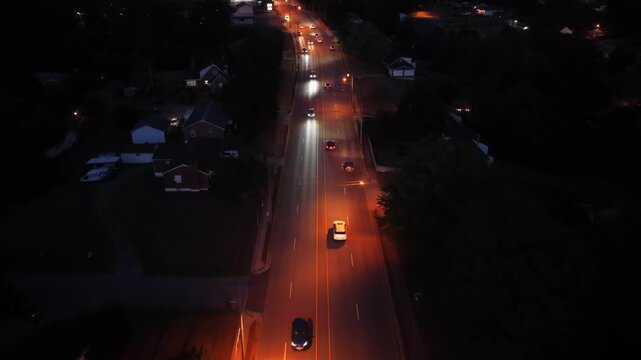 A drone flies forward over a lit arterial road in Lynchburg, Virginia, at night with two lines of traffic, street lamps and houses set back behind the trees. Aerial forward wide shot
