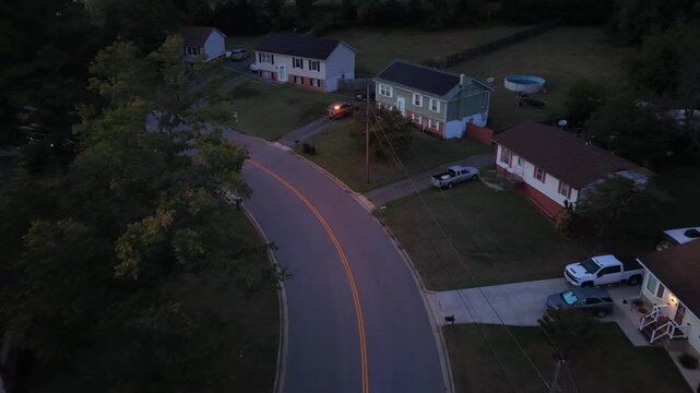 A curving suburban street in Lynchburg, Virginia, at dusk with split level homes, pickup trucks in the driveways, an above ground pool and mown lawns. Aerial lateral wide shot