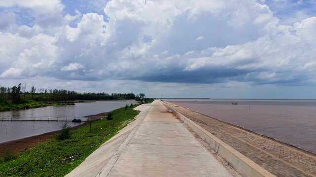 Visitors walk along the path at Con Nhan Beach, enjoying the view of the water and sky in Vinh Long, Vietnam. The scene shows boats in the distance and green areas along the shore.
