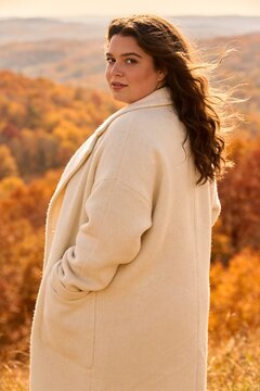 A woman in a cream wool coat stands on a windswept autumn hillside overlooking a valley of golden and rust colored trees, chubby girl.