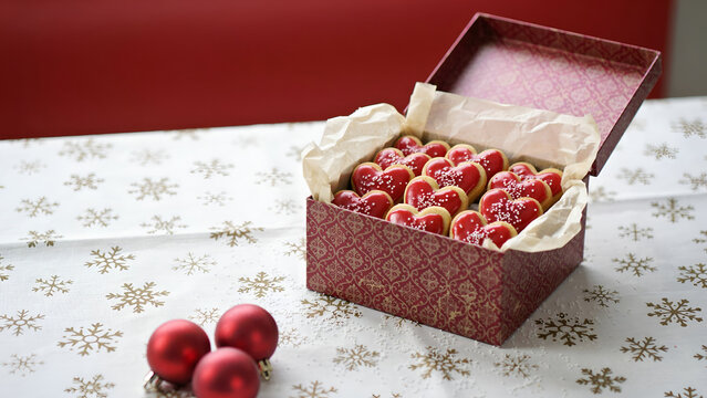 Heartshaped cookies with red icing and sprinkles in a decorative gift box on a snowflake patterned tablecloth with red ornaments