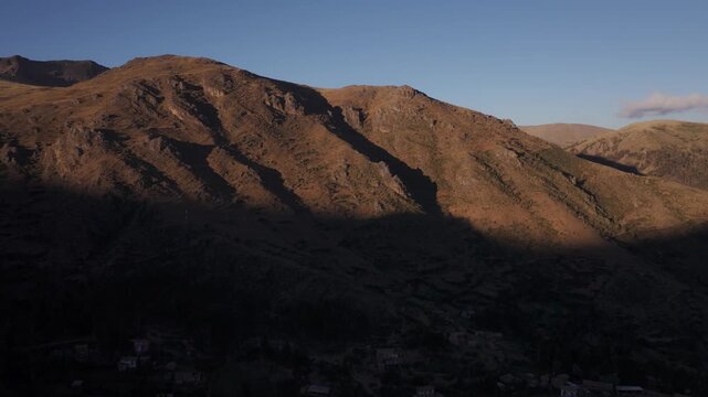 Aerial view of golden afternoon sunlight casting sharp shadows across rugged mountain ridges and arid slopes in the Andean highlands of Cusco region, Peru.