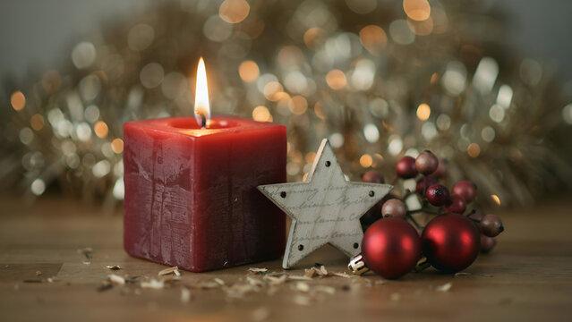 Christmas composition with a burning red square candle, a wooden star, and red ornaments on a wooden table with festive golden bokeh background