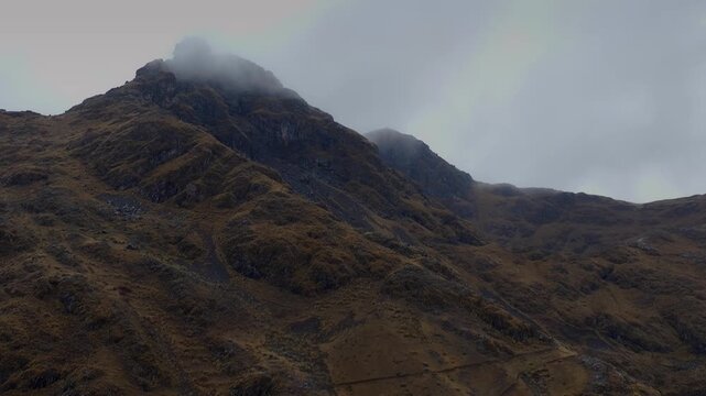 Cinematic aerial view of dramatic, rugged Andean mountain peaks shrouded in low hanging clouds and dense mist in the high-altitude wilderness of Cusco, Peru.