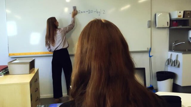 Over the shoulder view of a student attending a lesson while a teacher erases a math equation from a whiteboard in a modern classroom, with a laptop running code on the desk