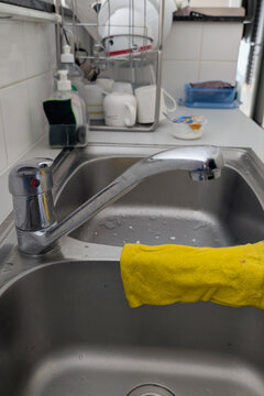 Hand with yellow cloth wipes kitchen sink area with faucet running during cleaning task