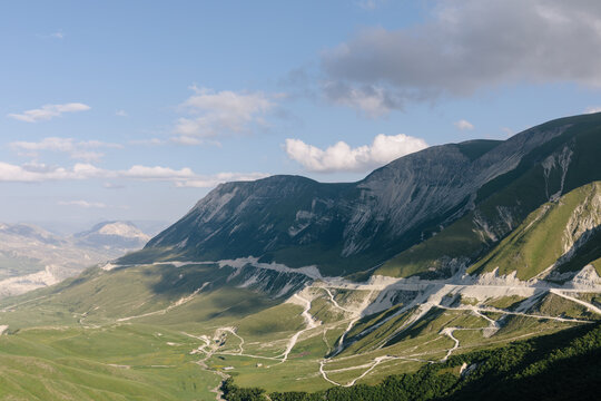 Wide panoramic view of the Matlas plateau with rugged mountain ridges, green valleys and winding dirt roads under a blue sky with scattered clouds