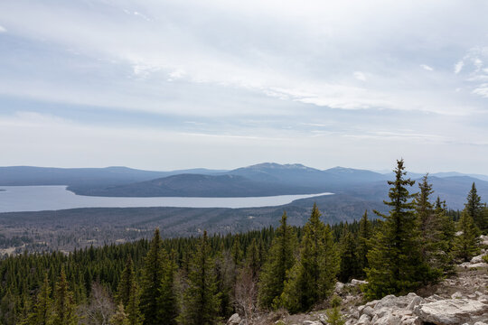 Wide landscape panorama of Zyuratkul National Park with lake shoreline, taiga conifer forest and distant Ural Mountains under cloudy sky, Russia