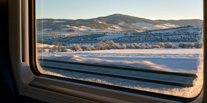 View of a winter landscape through a train window, with snow-covered fields, trees, and distant mountains under a clear sky