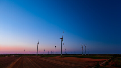 Obraz premium Wind turbines in field at blue hour. Dark generators stand in a wide rural landscape under a clear purple sky at dusk.