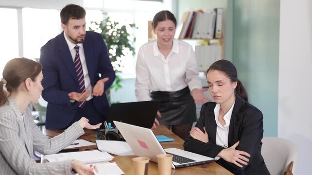 Group of women and man scold young woman while she sits looking sad in office 