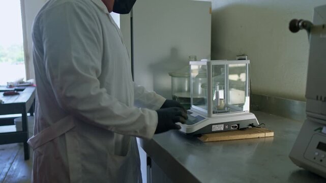 A lab technician in a white coat, gloves, and face mask carefully places or removes an item from a precision balance in a scientific laboratory. This work is part of a circular agribusiness and sustai