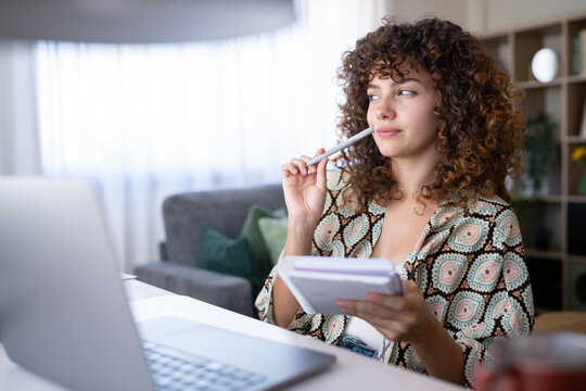 White woman at home holding a notebook and pen, thinking