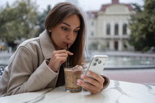 Young woman drinking iced coffee with straw and looking at smartphone at outdoor cafe terrace. Modern lifestyle, authentic emotion, casual tech concept