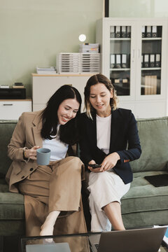 Two smiling women are comfortably sitting on a couch, sharing a moment and collaborating on a tablet, working together in a home office environment