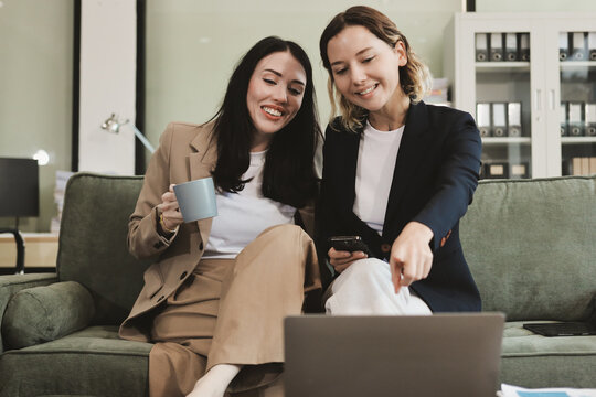 Two smiling women are comfortably sitting on a couch, sharing a moment and collaborating on a tablet, working together in a home office environment