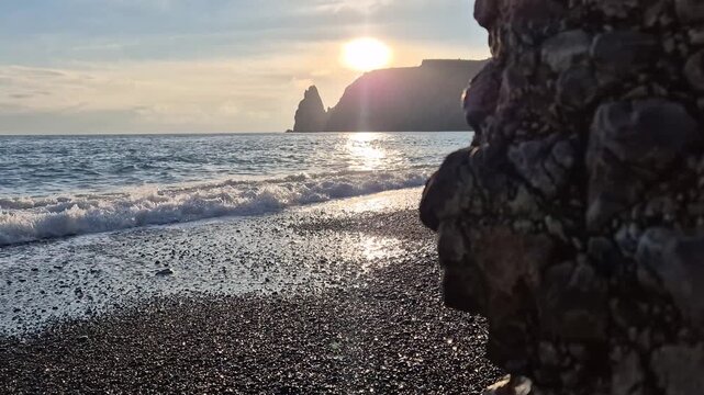 Ocean waves crash against a scenic rocky beach under a glowing sunset. Static ground-level shot.