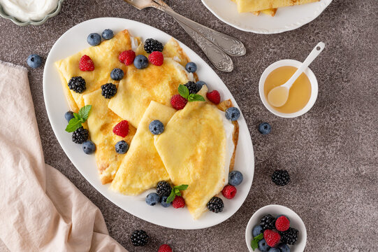 Crepes or thin pancakes with cream cheese, fresh berries and honey on a white plate on a concrete background. Copy space